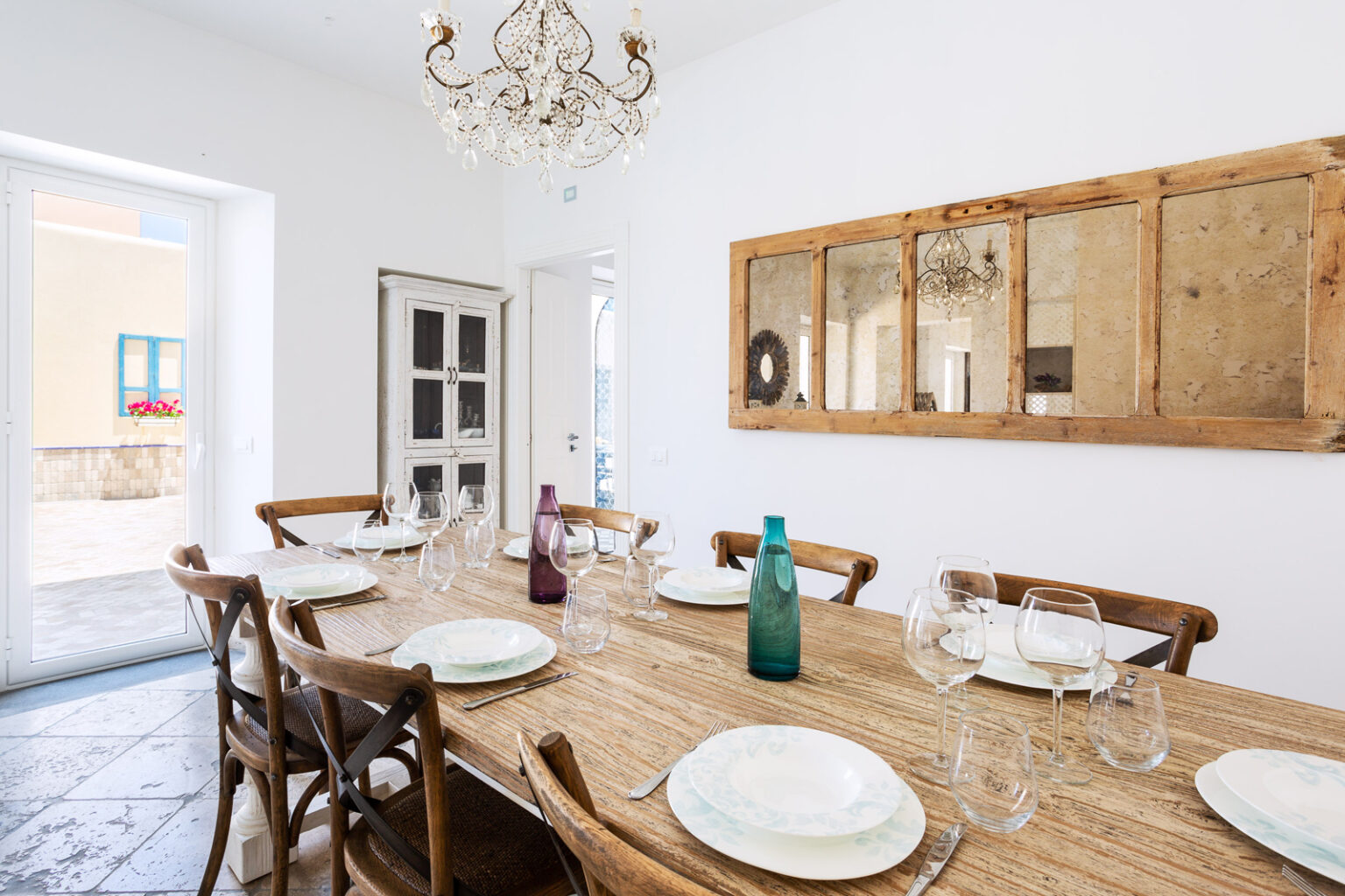 Elegant dining room featuring a rustic wooden table set for a meal, adorned with fine china and glassware. A stylish chandelier hangs from the ceiling, complementing the light-filled space with large windows and a view of outdoor flowers. The decor includes a vintage cabinet and a decorative mirror, enhancing the inviting atmosphere. | Only on SopranoVillas