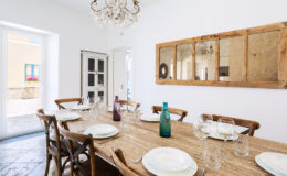 Elegant dining room featuring a rustic wooden table set for a meal, adorned with fine china and glassware. A stylish chandelier hangs from the ceiling, complementing the light-filled space with large windows and a view of outdoor flowers. The decor includes a vintage cabinet and a decorative mirror, enhancing the inviting atmosphere.