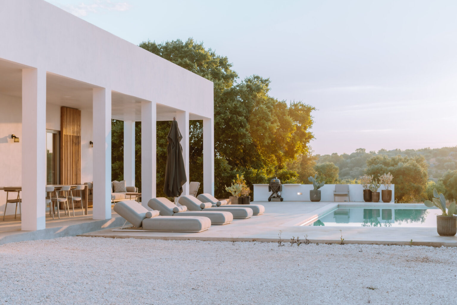 Modern villa exterior with a swimming pool and lounge chairs, surrounded by lush greenery and a serene landscape at sunset. The design features clean lines and a spacious terrace, ideal for relaxation and outdoor living. | Only on SopranoVillas
