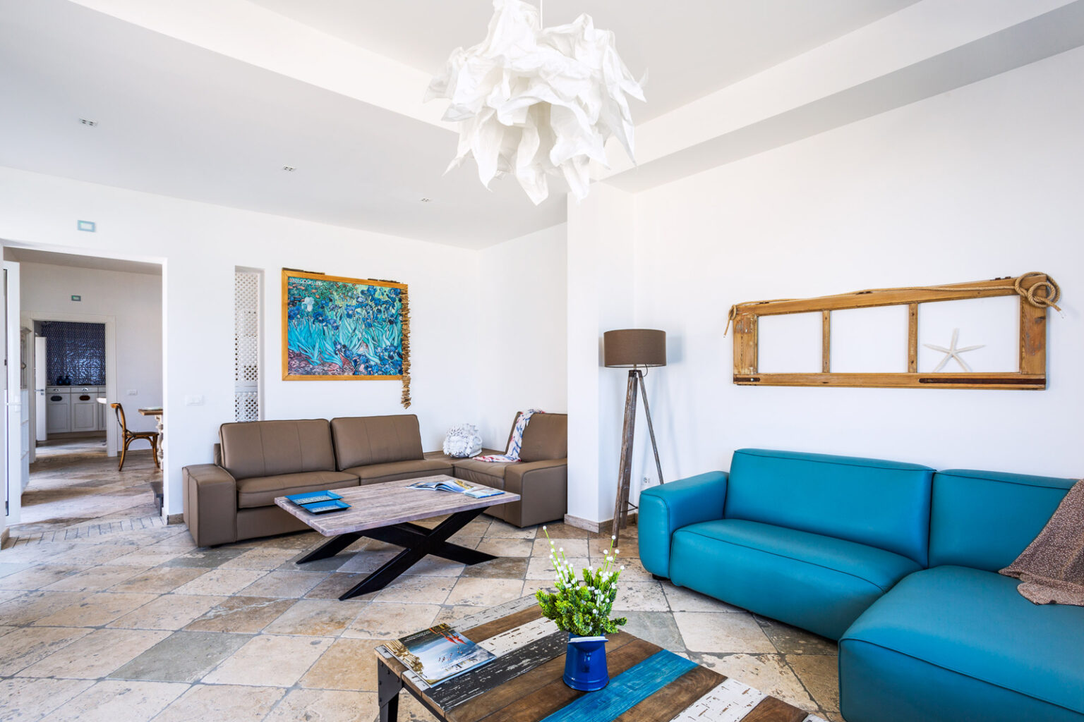 Modern living room interior featuring a brown sofa, a blue couch, and a stylish wooden coffee table. The room is adorned with a colorful piece of artwork, a unique light fixture, and decorative accents, creating a bright and inviting atmosphere. Natural stone flooring complements the contemporary design. | Only on SopranoVillas