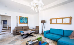 Modern living room interior featuring a brown sofa, a blue couch, and a stylish wooden coffee table. The room is adorned with a colorful piece of artwork, a unique light fixture, and decorative accents, creating a bright and inviting atmosphere. Natural stone flooring complements the contemporary design.