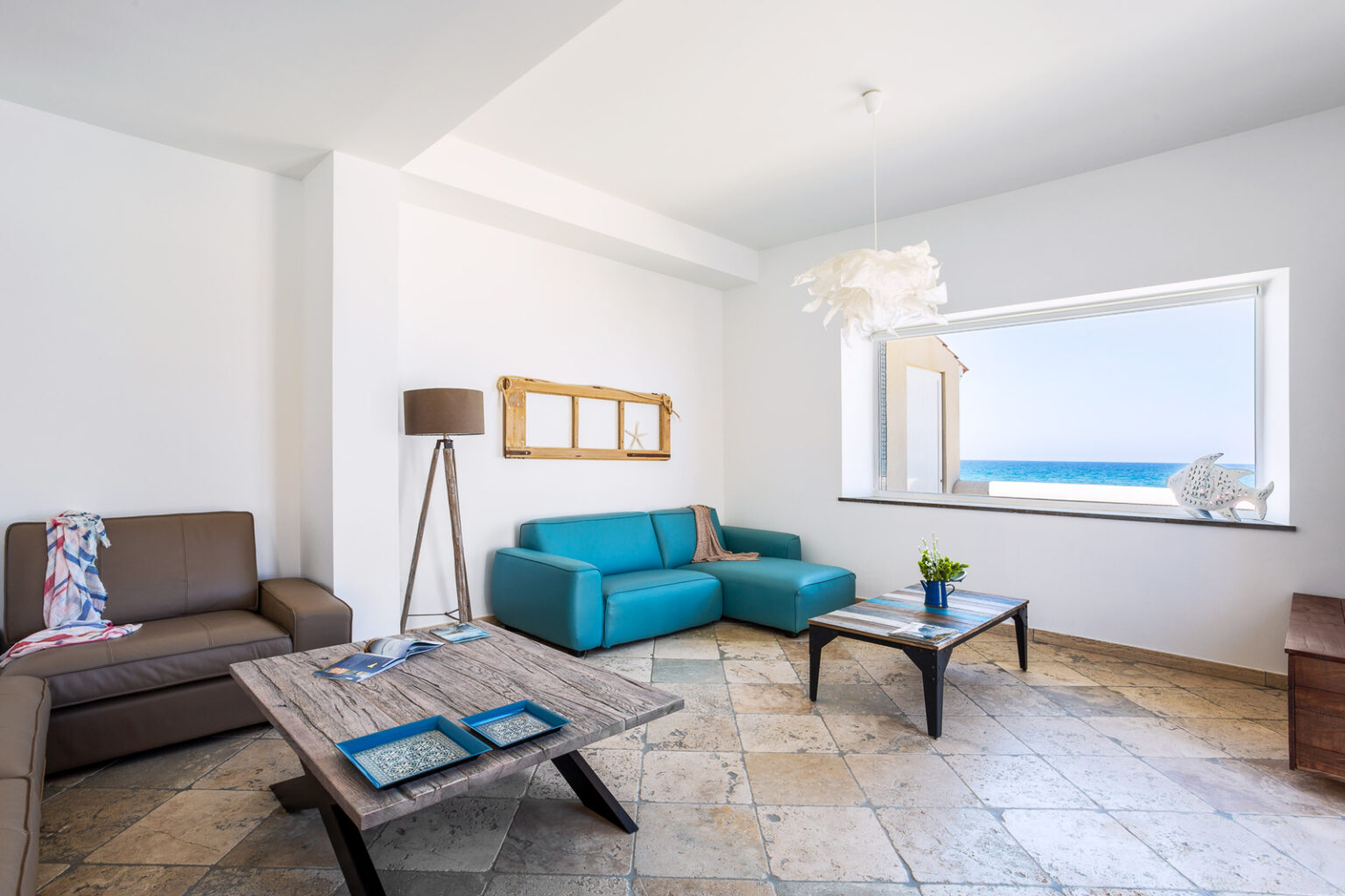 Modern coastal living room with a turquoise sofa, a brown leather couch, and a wooden coffee table. Large window showcasing a sea view, complemented by a stylish floor lamp and decorative elements. | Only on SopranoVillas