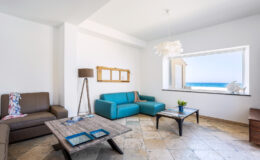 Modern coastal living room with a turquoise sofa, a brown leather couch, and a wooden coffee table. Large window showcasing a sea view, complemented by a stylish floor lamp and decorative elements.