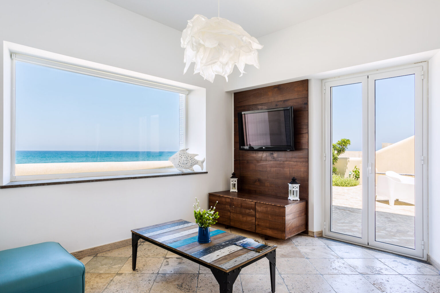 Modern coastal living room with large window showcasing ocean view, stylish furniture, and a flat-screen TV. Bright natural light enhances the serene atmosphere, perfect for relaxation. | Only on SopranoVillas