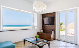 Modern coastal living room with large window showcasing ocean view, stylish furniture, and a flat-screen TV. Bright natural light enhances the serene atmosphere, perfect for relaxation.