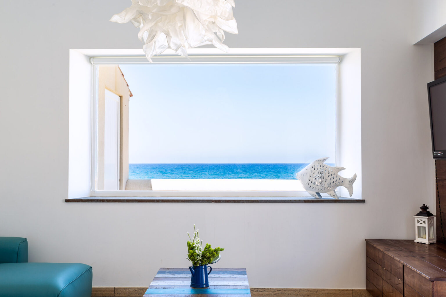 Bright coastal living room with a large window showcasing a stunning ocean view, featuring a blue sofa, decorative seashell, and a vase of fresh flowers on a coffee table. | Only on SopranoVillas