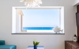 Bright coastal living room with a large window showcasing a stunning ocean view, featuring a blue sofa, decorative seashell, and a vase of fresh flowers on a coffee table.
