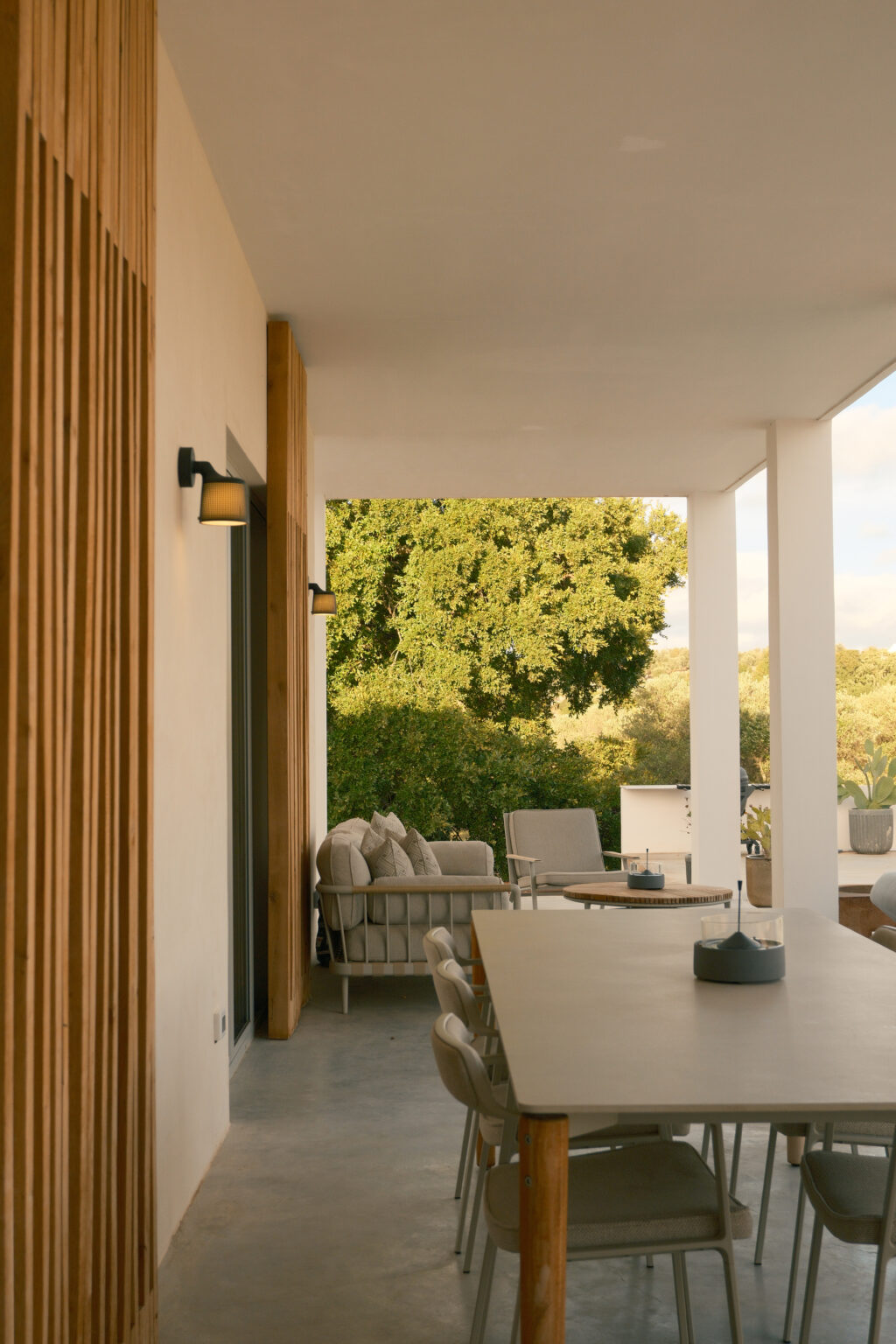 Modern outdoor patio featuring a spacious dining table, stylish chairs, and comfortable seating area with cushions. The design incorporates wooden accents and ambient lighting, surrounded by lush greenery for a serene atmosphere. | Only on SopranoVillas
