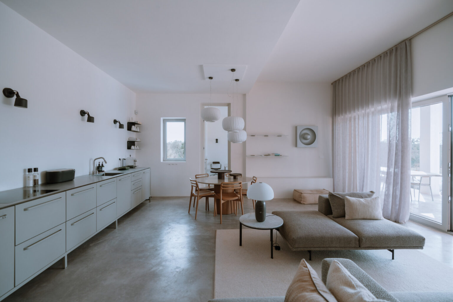 Modern open-concept kitchen and living room featuring minimalist design, light-colored cabinetry, a cozy sofa, and a dining area with wooden furniture. Large windows allow natural light to flood the space, enhancing its airy atmosphere. | Only on SopranoVillas