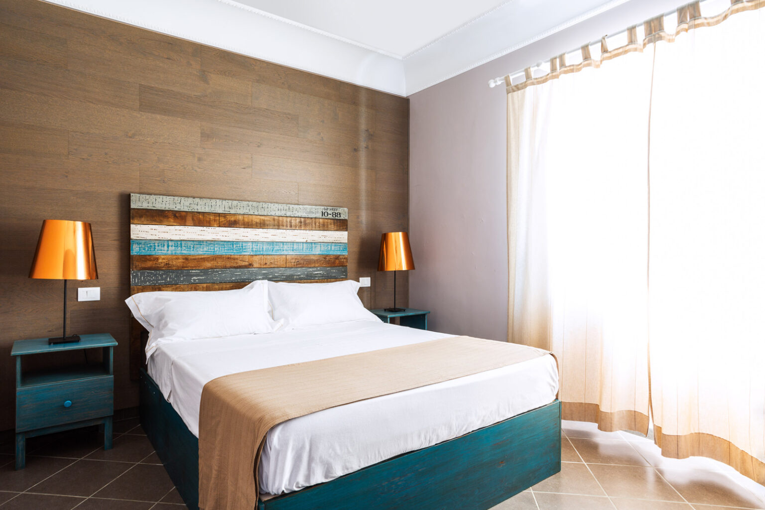 Cozy modern bedroom featuring a queen-size bed with a beige and white comforter, wooden accent wall, turquoise nightstands, and stylish orange lamps, illuminated by natural light from a large window with sheer curtains. | Only on SopranoVillas