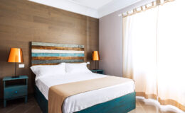 Cozy modern bedroom featuring a queen-size bed with a beige and white comforter, wooden accent wall, turquoise nightstands, and stylish orange lamps, illuminated by natural light from a large window with sheer curtains.