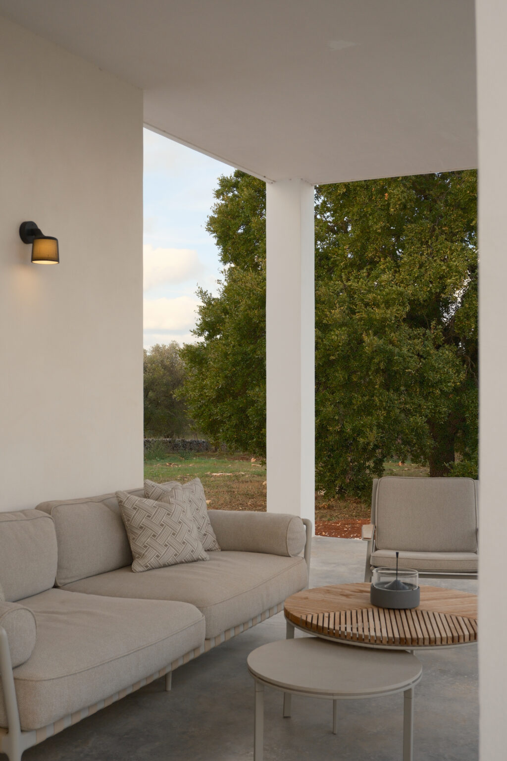 Modern outdoor seating area featuring a stylish beige sofa with decorative pillows, a round wooden coffee table, and a minimalist chair, set against a backdrop of greenery. Ideal for relaxation and outdoor entertaining. | Only on SopranoVillas