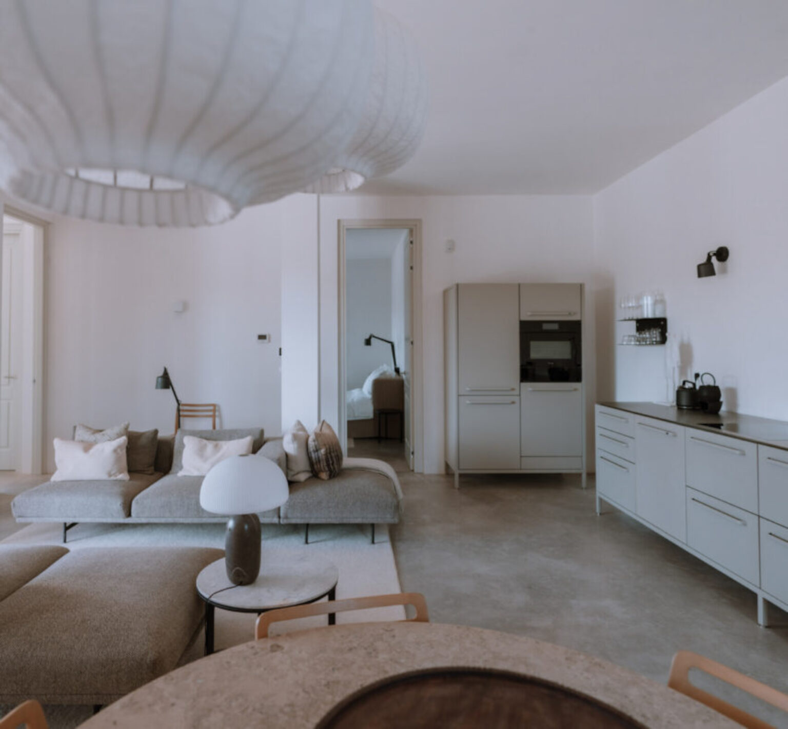 Modern living room interior featuring a sleek gray sofa with decorative pillows, a minimalist coffee table, and a stylish lamp. The room includes a contemporary kitchen with a built-in refrigerator and cabinetry, all set against a light-colored wall for a spacious feel. | Only on SopranoVillas