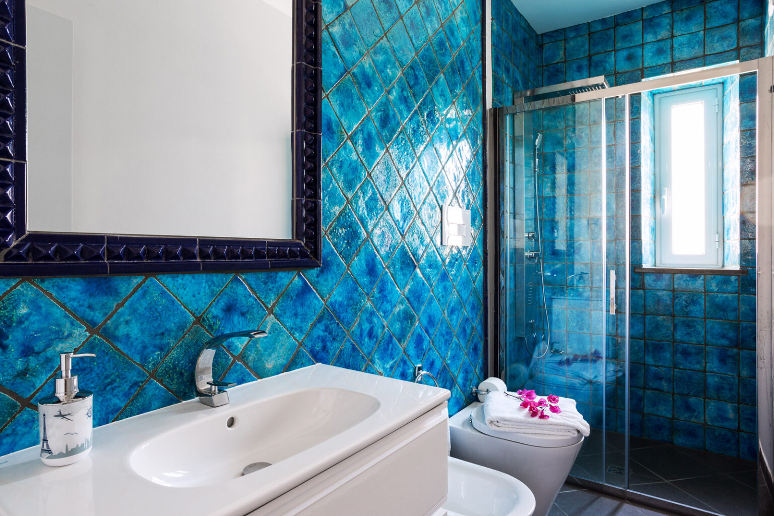 Modern bathroom featuring vibrant blue tiled walls, a sleek white vanity with a sink, and a glass shower enclosure. The decor includes a stylish mirror and a touch of floral accents on the towels, creating a refreshing and inviting atmosphere. | Only on SopranoVillas