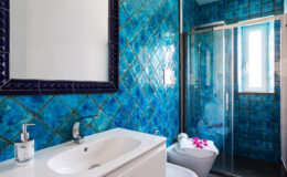 Modern bathroom featuring vibrant blue tiled walls, a sleek white vanity with a sink, and a glass shower enclosure. The decor includes a stylish mirror and a touch of floral accents on the towels, creating a refreshing and inviting atmosphere.