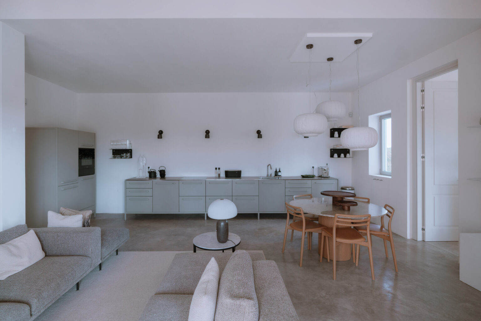 Modern open-concept living room and kitchen featuring minimalist design, light gray furniture, wooden dining table, and contemporary lighting fixtures. | Only on SopranoVillas