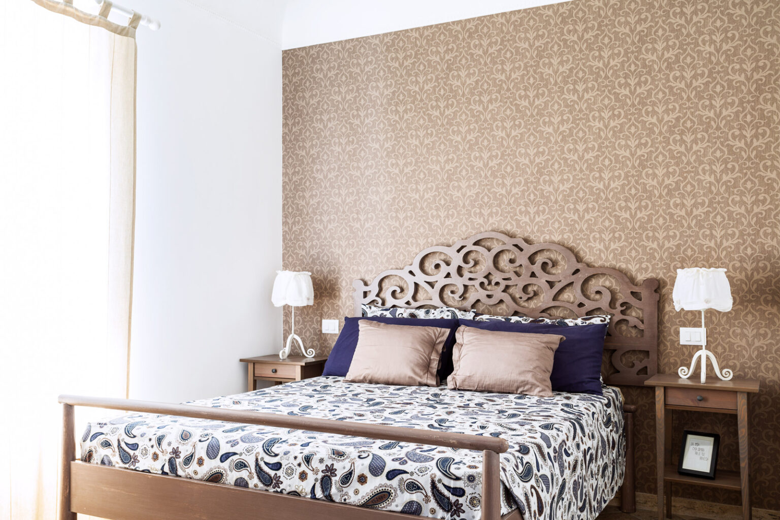 Cozy bedroom featuring a wooden bed frame with an ornate headboard, decorated with patterned bedding and plush pillows. Two bedside tables with stylish lamps complement the warm, textured wall design. Natural light filters through a window, enhancing the inviting atmosphere. | Only on SopranoVillas