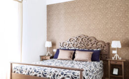 Cozy bedroom featuring a wooden bed frame with an ornate headboard, decorated with patterned bedding and plush pillows. Two bedside tables with stylish lamps complement the warm, textured wall design. Natural light filters through a window, enhancing the inviting atmosphere.
