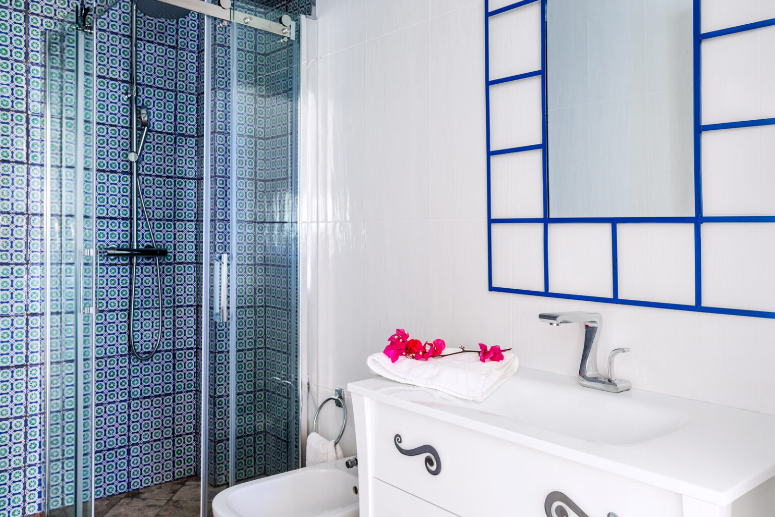 Modern bathroom featuring a glass shower, decorative blue tiled wall, white sink with a stylish faucet, and pink flower accents on the counter. | Only on SopranoVillas