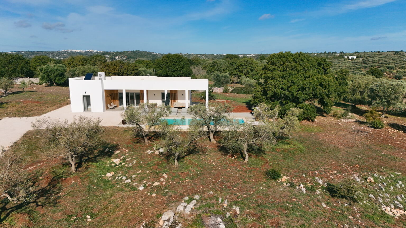 Aerial view of a modern white villa with a swimming pool surrounded by olive trees and lush greenery, set in a tranquil landscape under a clear blue sky. | Only on SopranoVillas