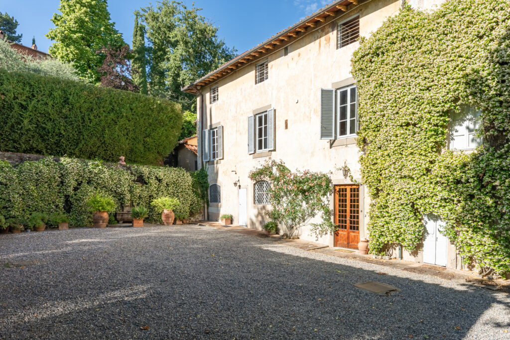 Historic villa surrounded by lush greenery and flowering plants, featuring a gravel courtyard, large windows, and rustic architecture, ideal for outdoor gatherings and relaxation. | Only on SopranoVillas