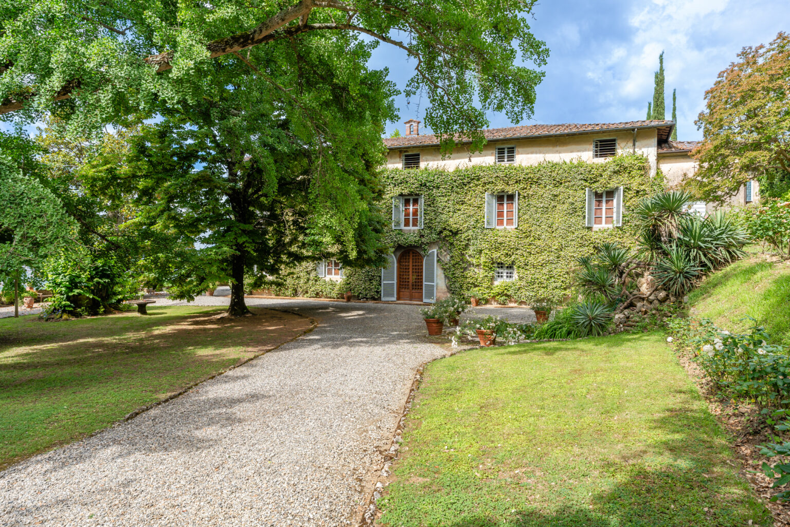 Charming Italian villa surrounded by lush greenery, featuring ivy-covered walls, a gravel pathway, and potted plants in a serene garden setting. Ideal for nature lovers and architecture enthusiasts. | Only on SopranoVillas