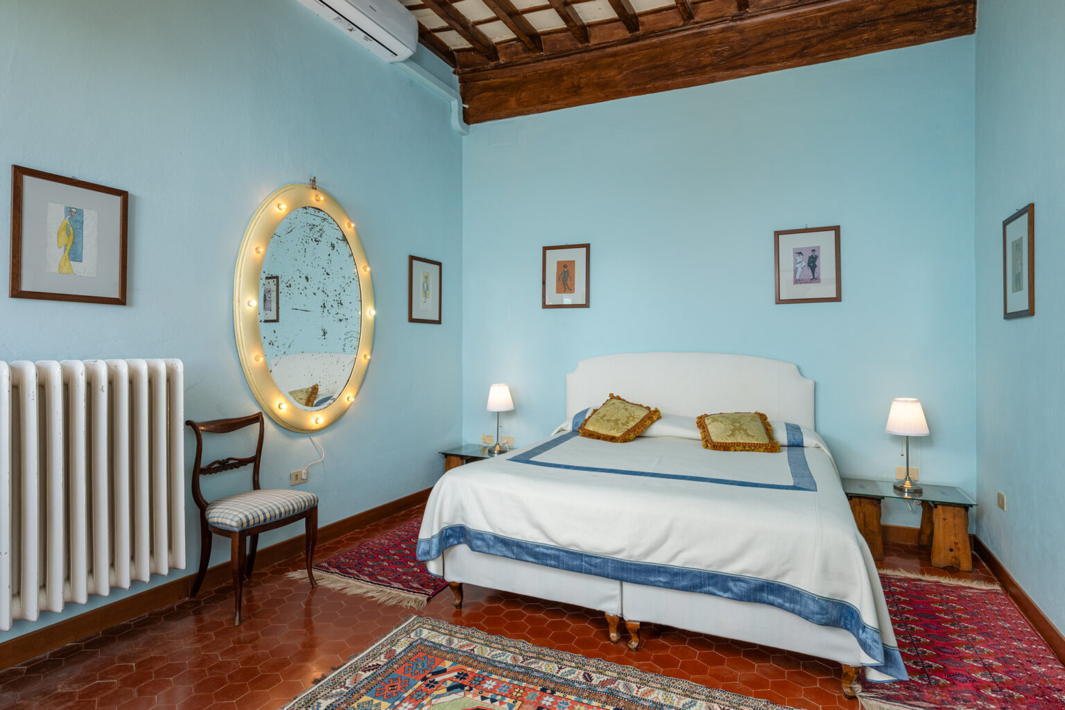 Cozy bedroom featuring a light blue color scheme, a double bed with decorative pillows, a vintage chair, a round mirror with lights, and framed artwork on the walls. The room has a warm ambiance with wooden ceiling beams and a patterned area rug. | Only on SopranoVillas