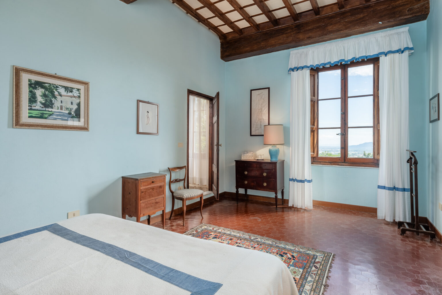 Bright and airy bedroom featuring light blue walls, wooden ceiling beams, and a large window with scenic views. The room includes a bed, a wooden dresser, a chair, and decorative artwork, creating a cozy and inviting atmosphere. | Only on SopranoVillas