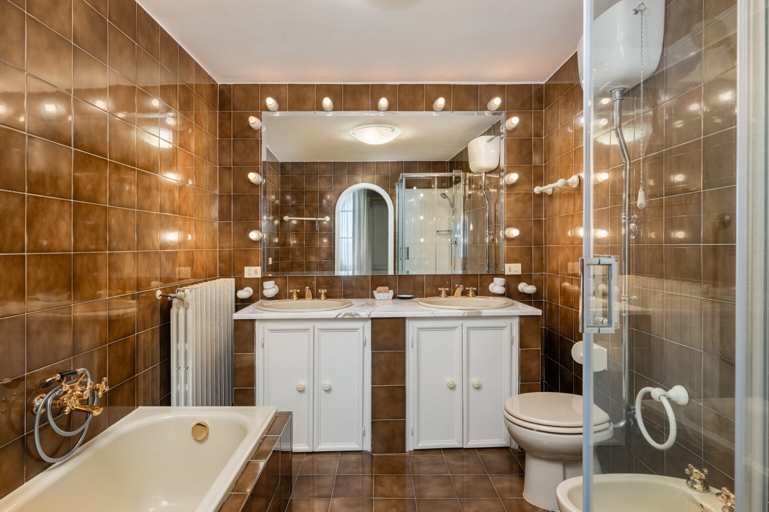 Modern bathroom featuring brown tiled walls, a double vanity with bright lights, a bathtub, a toilet, and a glass shower enclosure. Ideal for showcasing stylish and functional bathroom design. | Only on SopranoVillas