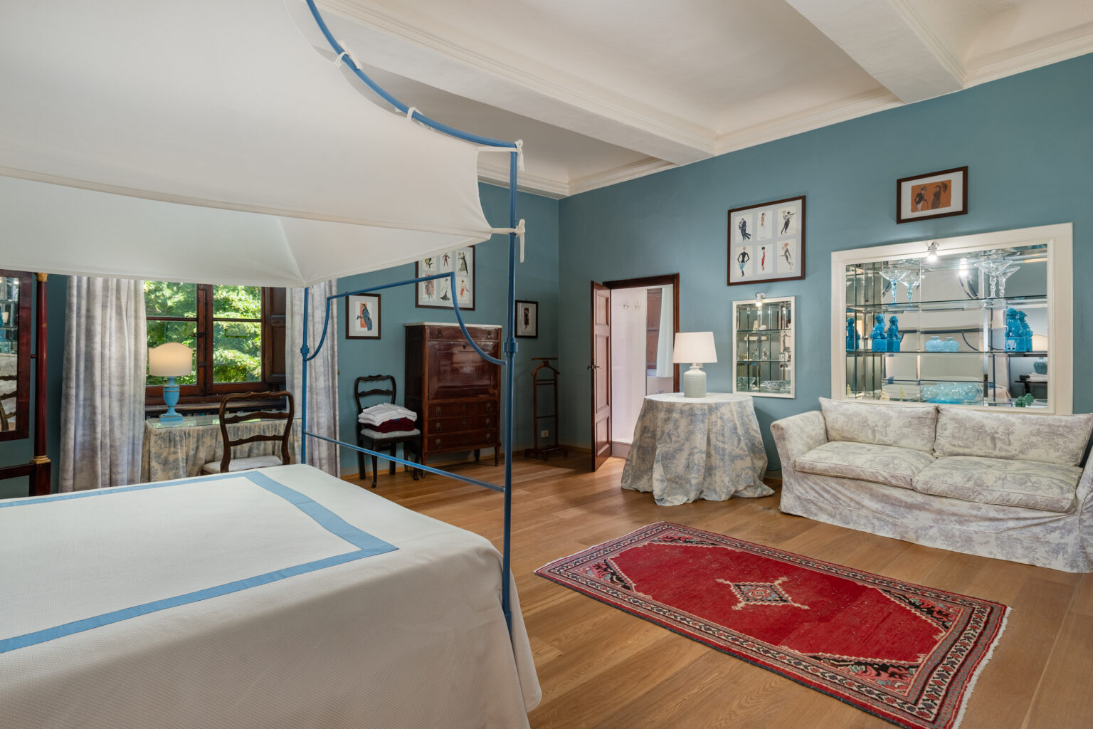 Spacious bedroom featuring a four-poster bed, elegant blue walls, and wooden flooring. The room includes a cozy seating area with a patterned rug, a dressing table, and framed artwork, creating a warm and inviting atmosphere. Natural light filters through the windows, enhancing the room's charm. | Only on SopranoVillas
