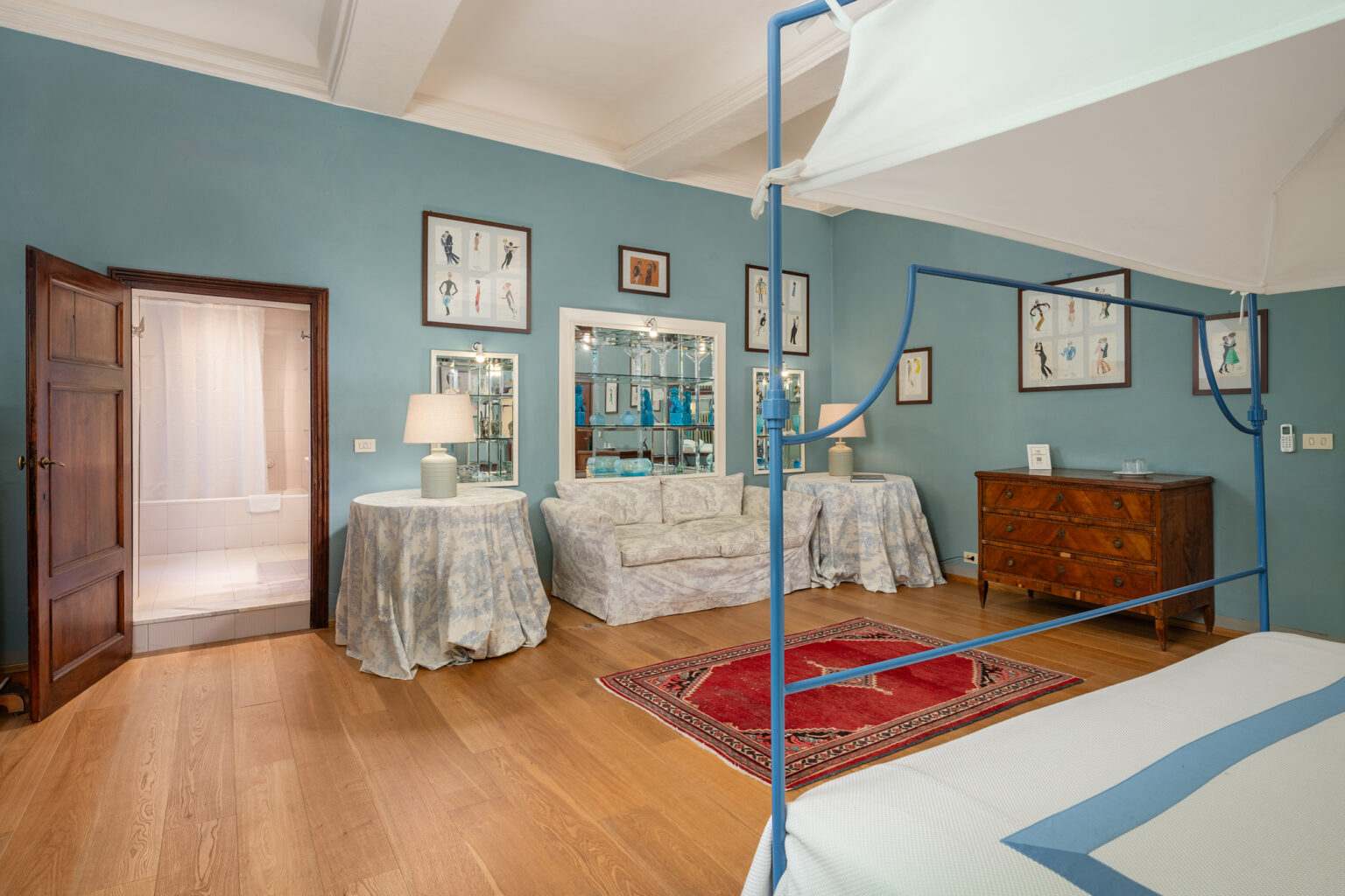 Spacious bedroom interior featuring a blue canopy bed, elegant wooden furniture, and decorative artwork on the walls. The room includes a cozy seating area with floral-patterned upholstery, a bedside lamp, and an adjoining bathroom visible through an open door. The warm wooden flooring and soft blue walls create a serene atmosphere. | Only on SopranoVillas