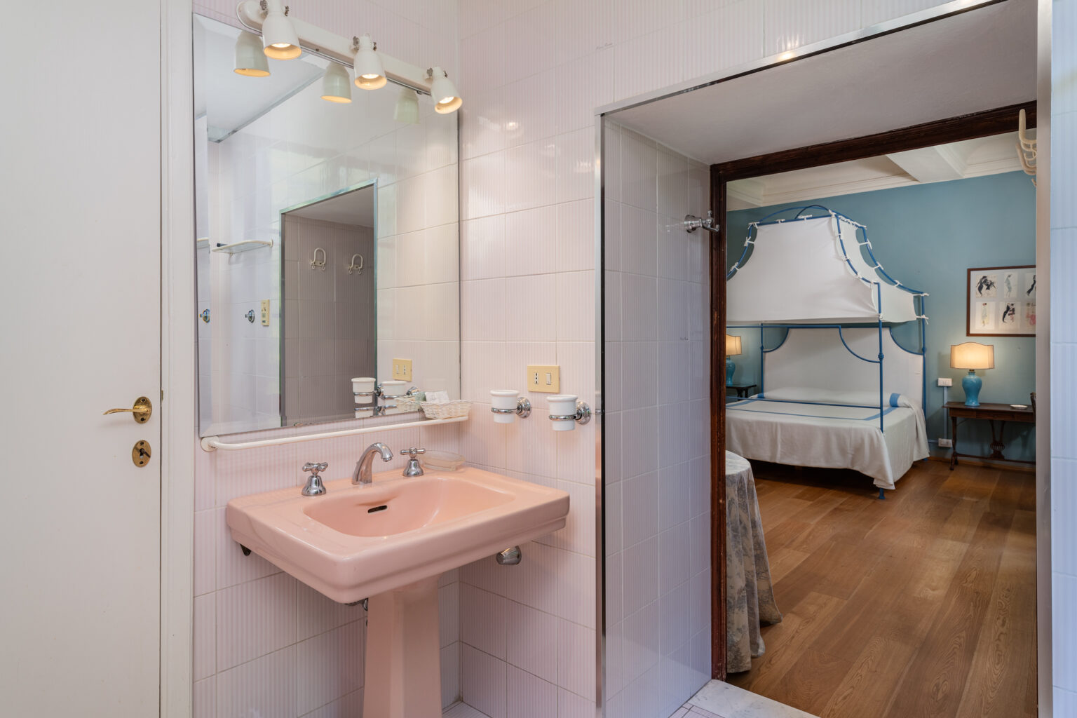 Modern bathroom with a pink sink and mirror, featuring a doorway that leads to a stylish bedroom with a four-poster bed and blue walls. | Only on SopranoVillas
