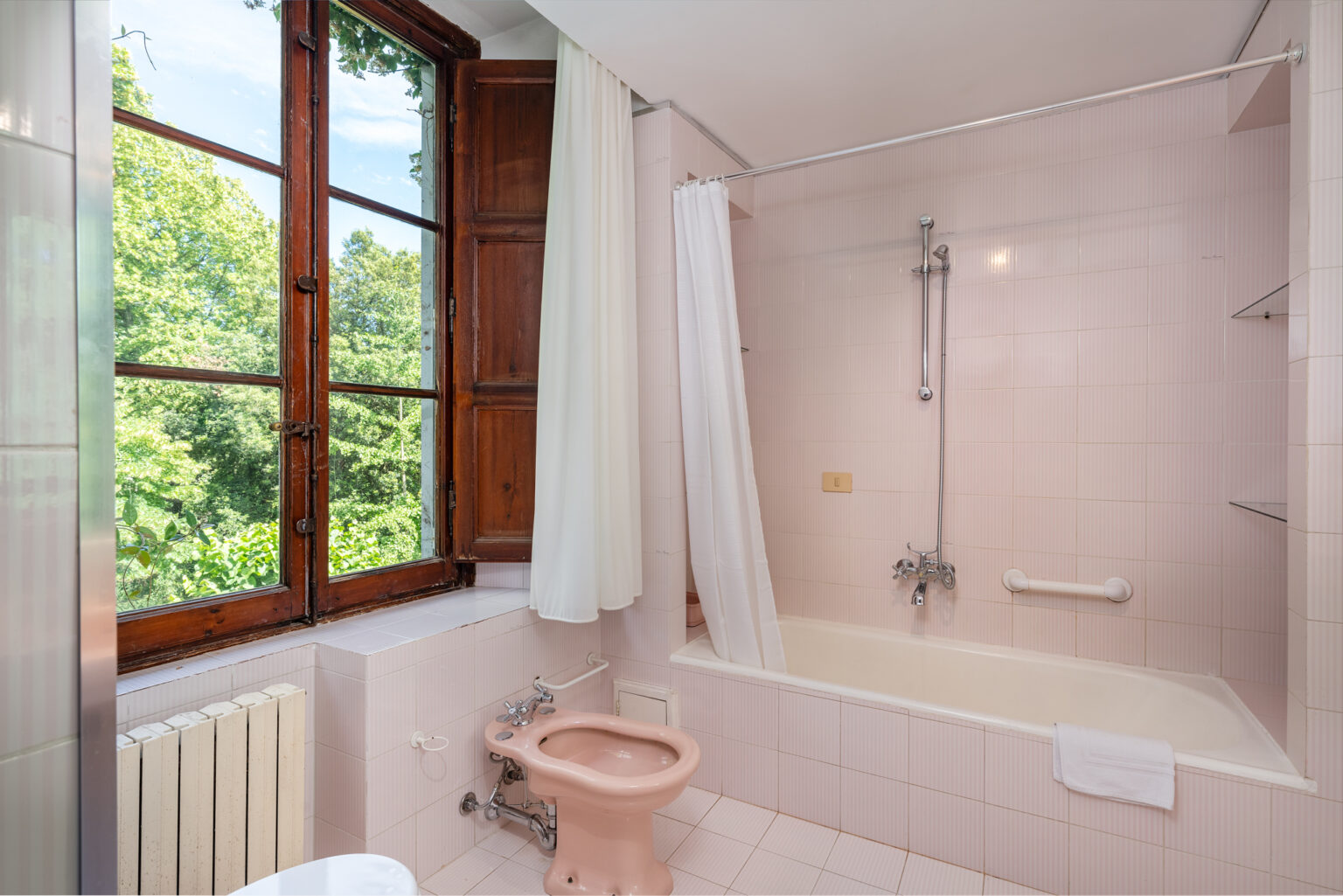 Bright and airy bathroom featuring a bathtub with shower, pink tiles, and large windows overlooking greenery. The design includes a toilet and a towel rack, creating a serene space for relaxation. | Only on SopranoVillas