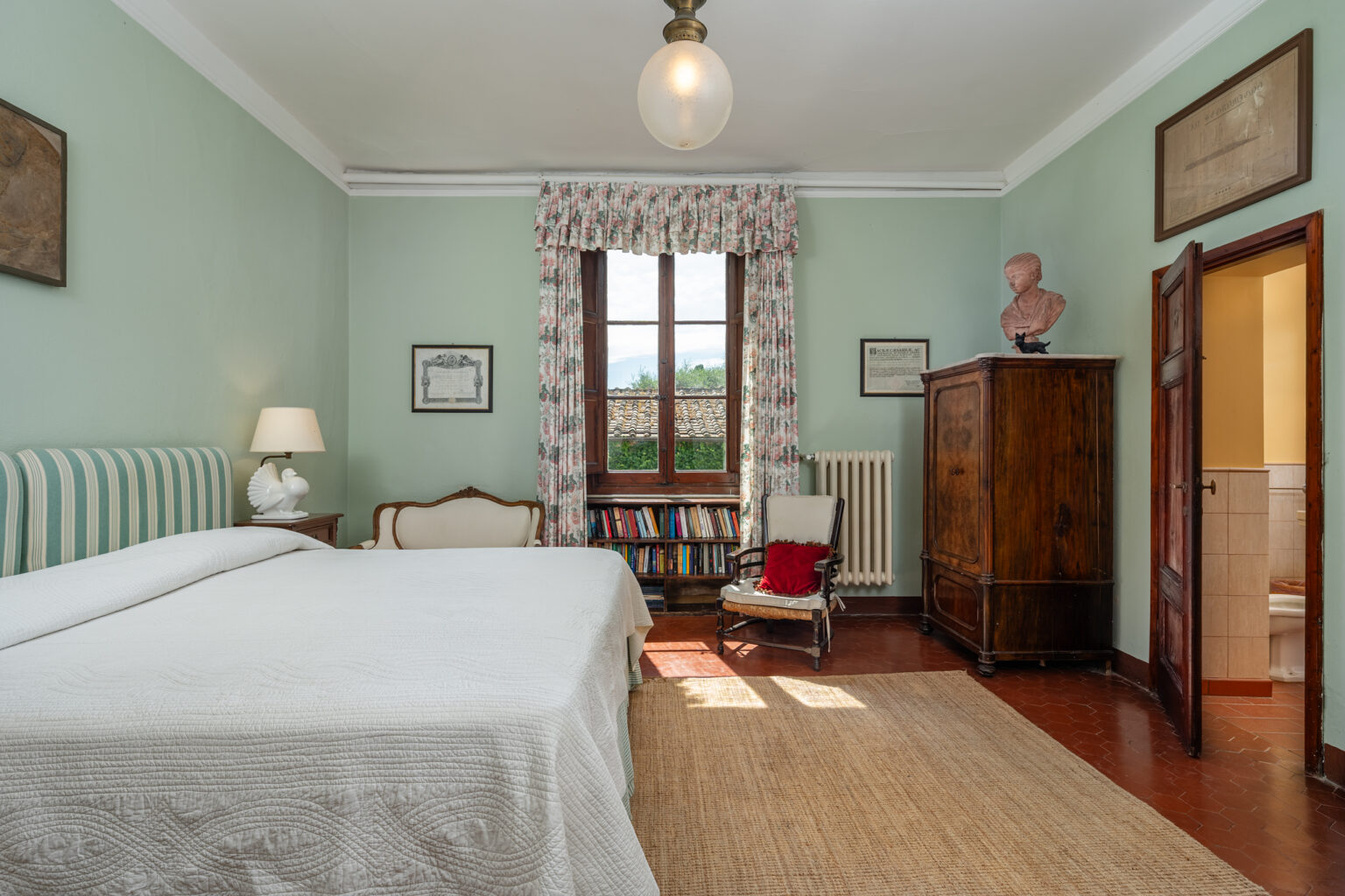Cozy bedroom with light green walls, featuring a double bed with white linens, a vintage wooden wardrobe, a small reading nook with a chair, and natural light streaming through large windows. Decorated with artwork and a bust sculpture for a classic touch. | Only on SopranoVillas