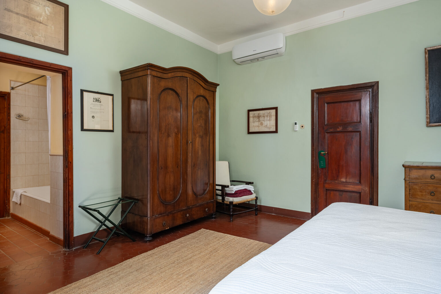 Cozy bedroom featuring a vintage wooden wardrobe, a small chair with folded towels, and a rustic rug. The walls are painted a soft green, complemented by wooden accents and framed artwork. An air conditioning unit is installed for comfort, and a doorway leads to a bathroom area. | Only on SopranoVillas