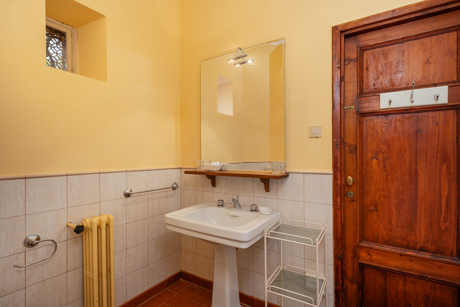 Bright and airy bathroom featuring a white sink, chrome fixtures, and a wooden door. The walls are painted yellow, complemented by light-colored tiles. A mirror with a light above it and a wooden shelf with toiletries enhance the space. A small window allows natural light, and a white shelving unit adds storage. | Only on SopranoVillas