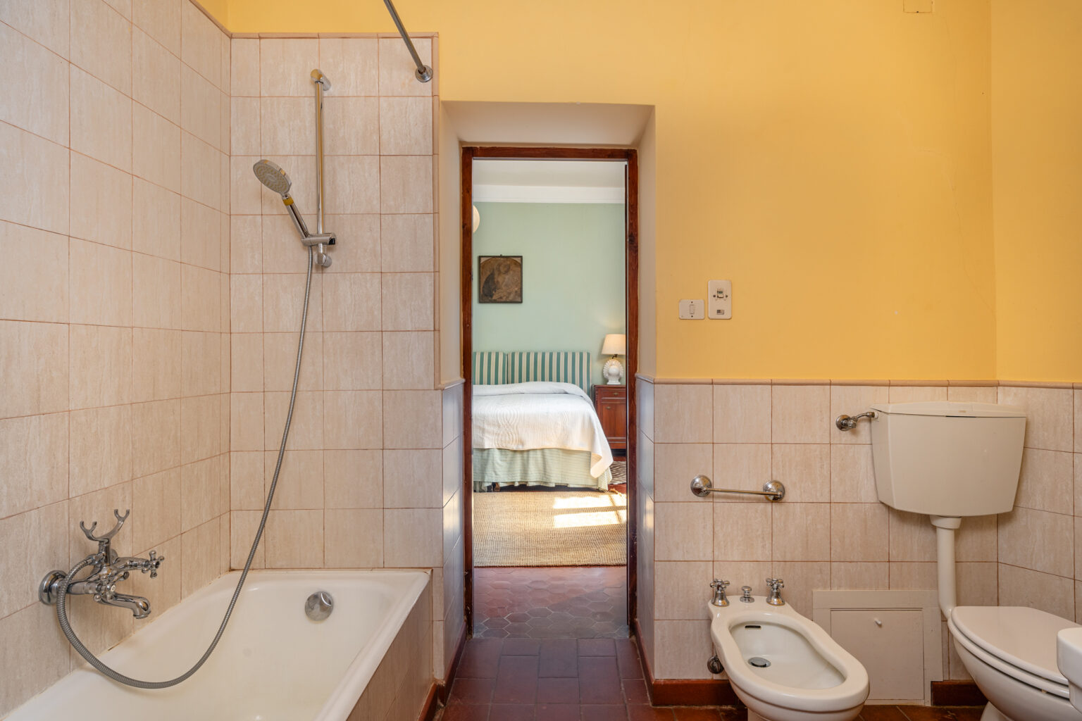 Modern bathroom featuring a bathtub with shower, toilet, and bidet, with a doorway leading to a well-lit bedroom. The walls are tiled in neutral tones, and the decor includes warm yellow and green accents. | Only on SopranoVillas