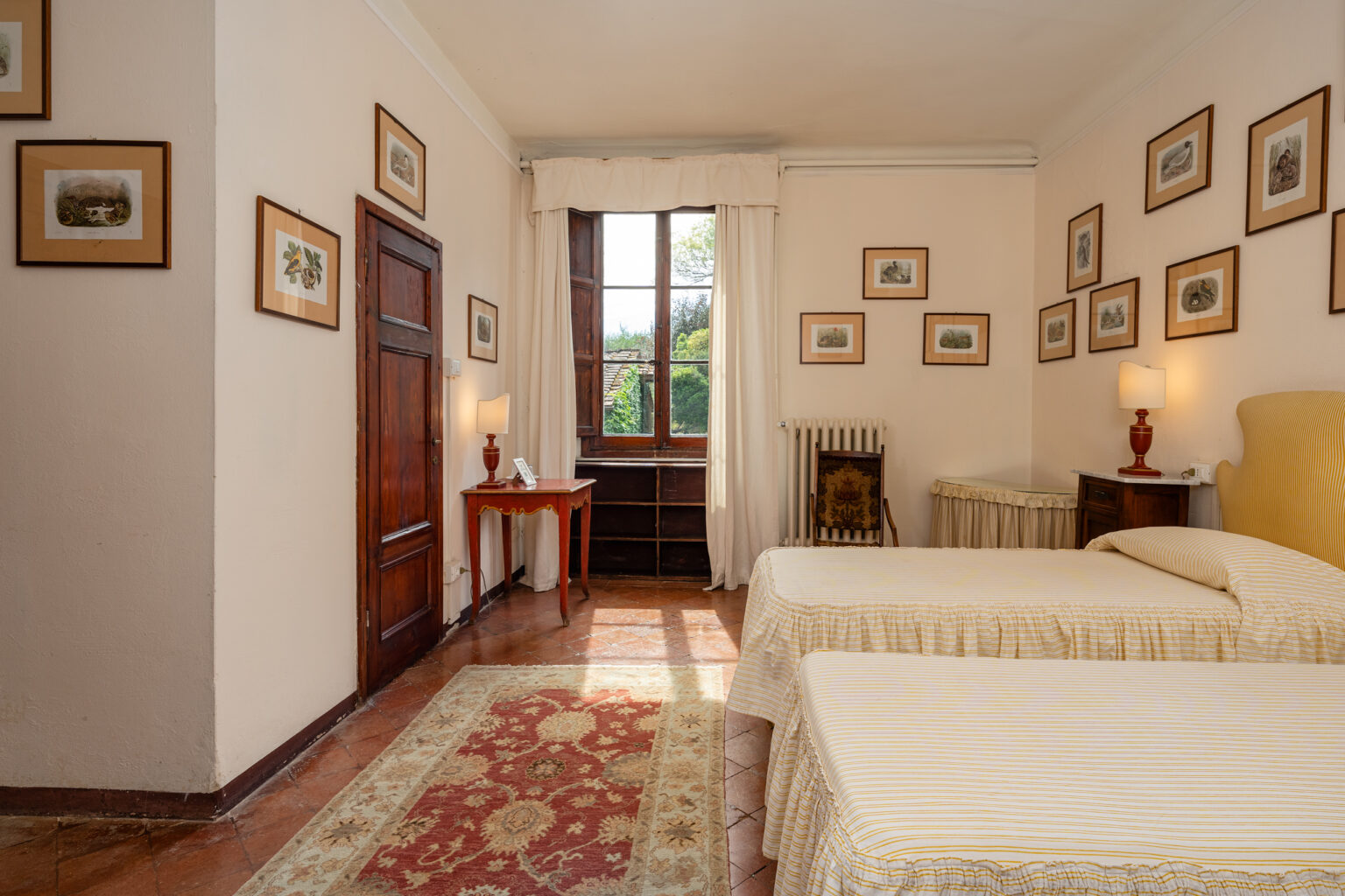 Cozy bedroom featuring two twin beds with yellow striped bedding, a wooden desk with a lamp, and framed artwork on the walls. Natural light streams through a large window, highlighting a warm and inviting atmosphere. | Only on SopranoVillas