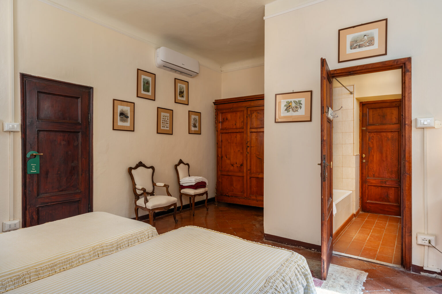 Cozy bedroom interior featuring two twin beds, vintage wooden furniture, and framed artwork on the walls. The room includes a wardrobe, seating area with chairs, and an ensuite bathroom, providing a comfortable and inviting atmosphere. | Only on SopranoVillas