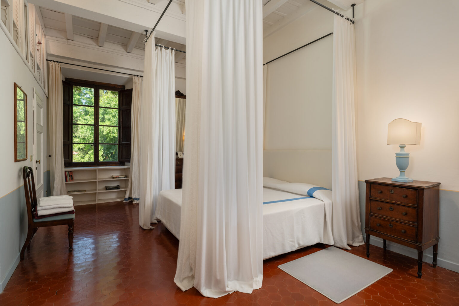 Cozy bedroom featuring a bed with white drapery, a wooden nightstand with a blue lamp, and large windows allowing natural light, set in a serene environment. | Only on SopranoVillas