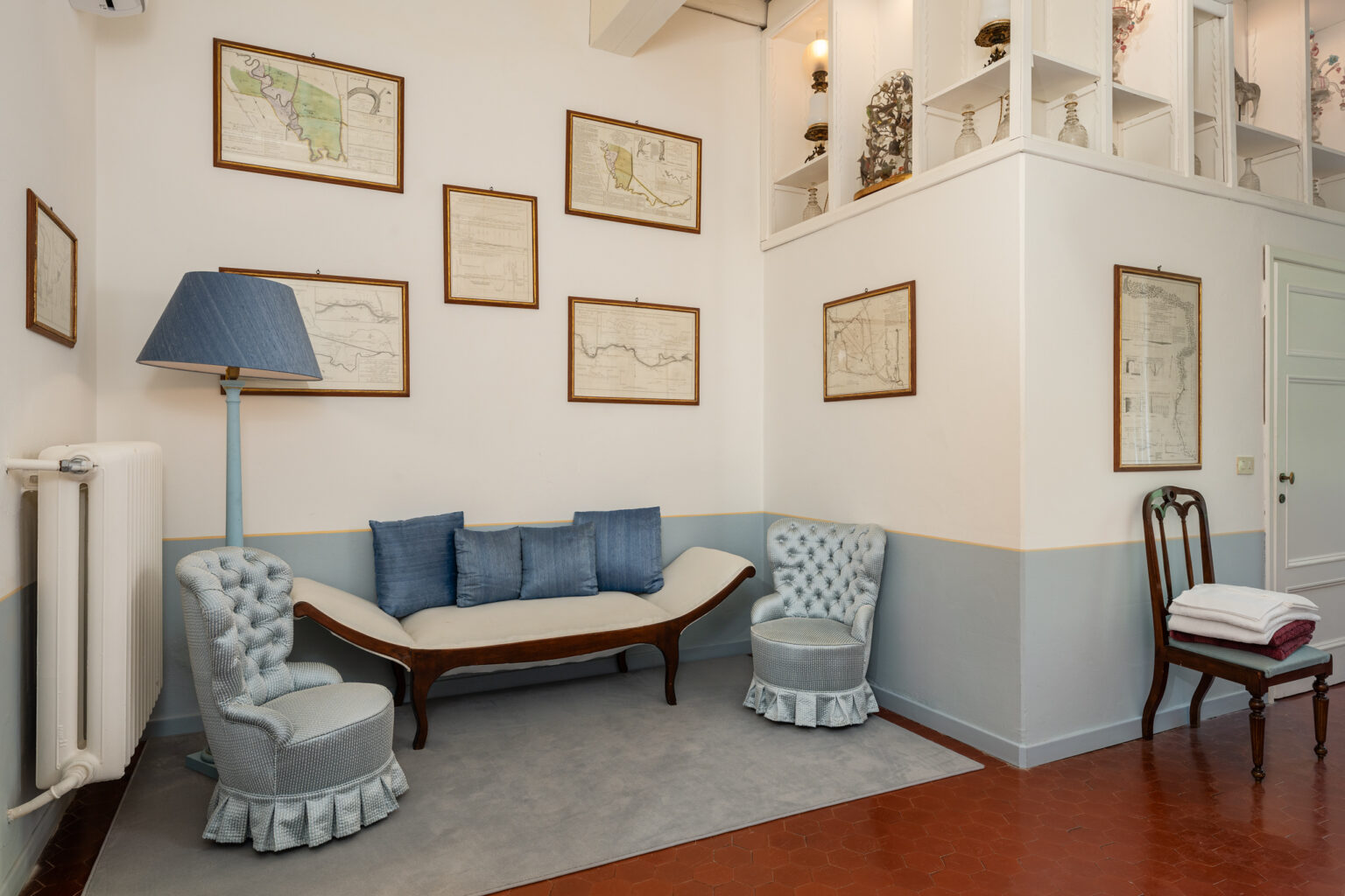 Cozy interior of a living room featuring vintage decor, including framed historical maps on the walls, a stylish light blue lamp, and elegant upholstered chairs. The space is complemented by a wooden settee with blue cushions and a neatly arranged wooden chair with folded towels. | Only on SopranoVillas