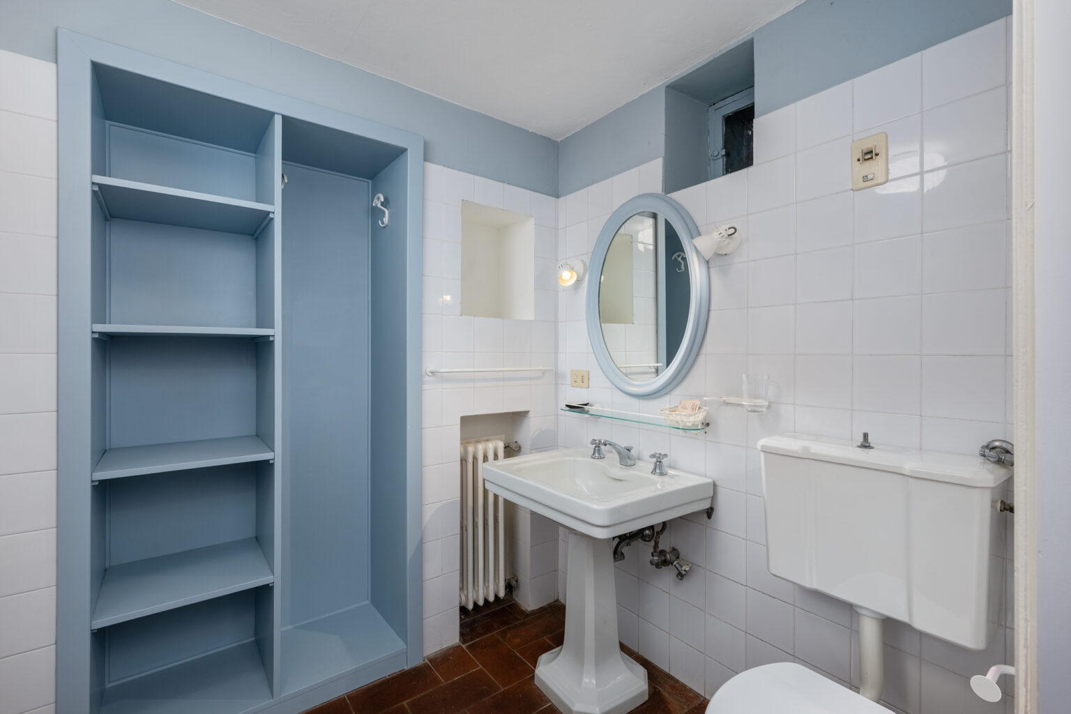 Bright and modern bathroom featuring a white pedestal sink, a toilet, and a round mirror with a blue frame. The walls are adorned with white tiles and light blue accents, complemented by built-in shelving for storage. Natural light streams in through a small window, enhancing the airy atmosphere. | Only on SopranoVillas