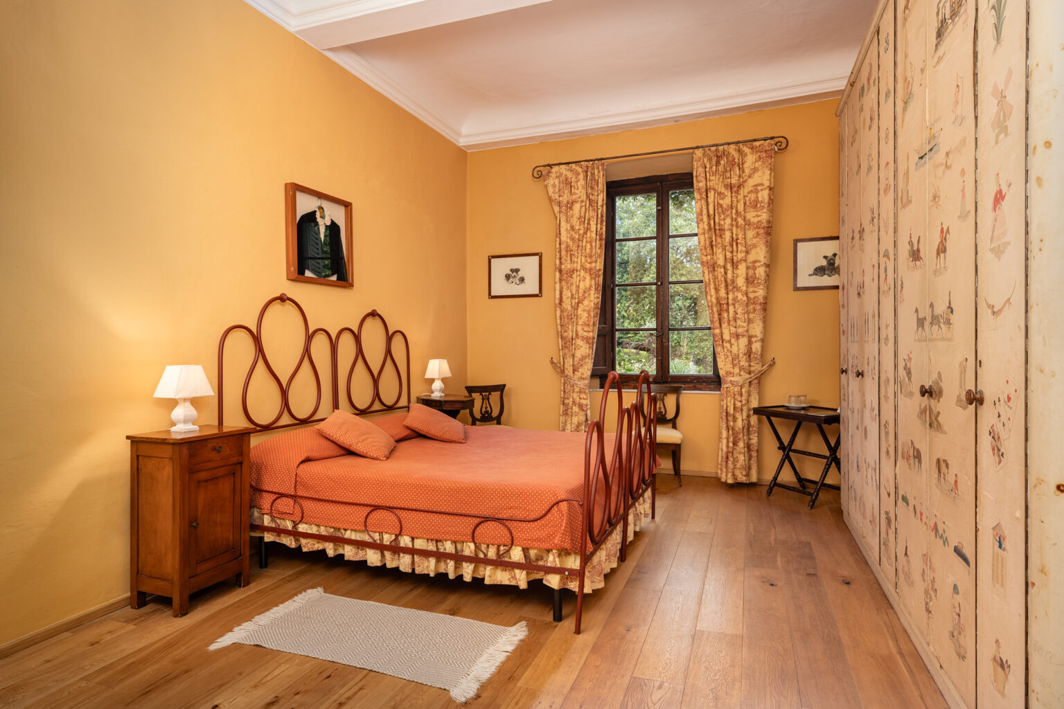 Cozy bedroom featuring a vintage red metal bed frame with orange bedding, complemented by wooden furniture, decorative curtains, and a charming wardrobe adorned with illustrations. Natural light filters through a large window, enhancing the warm ambiance of the space. | Only on SopranoVillas