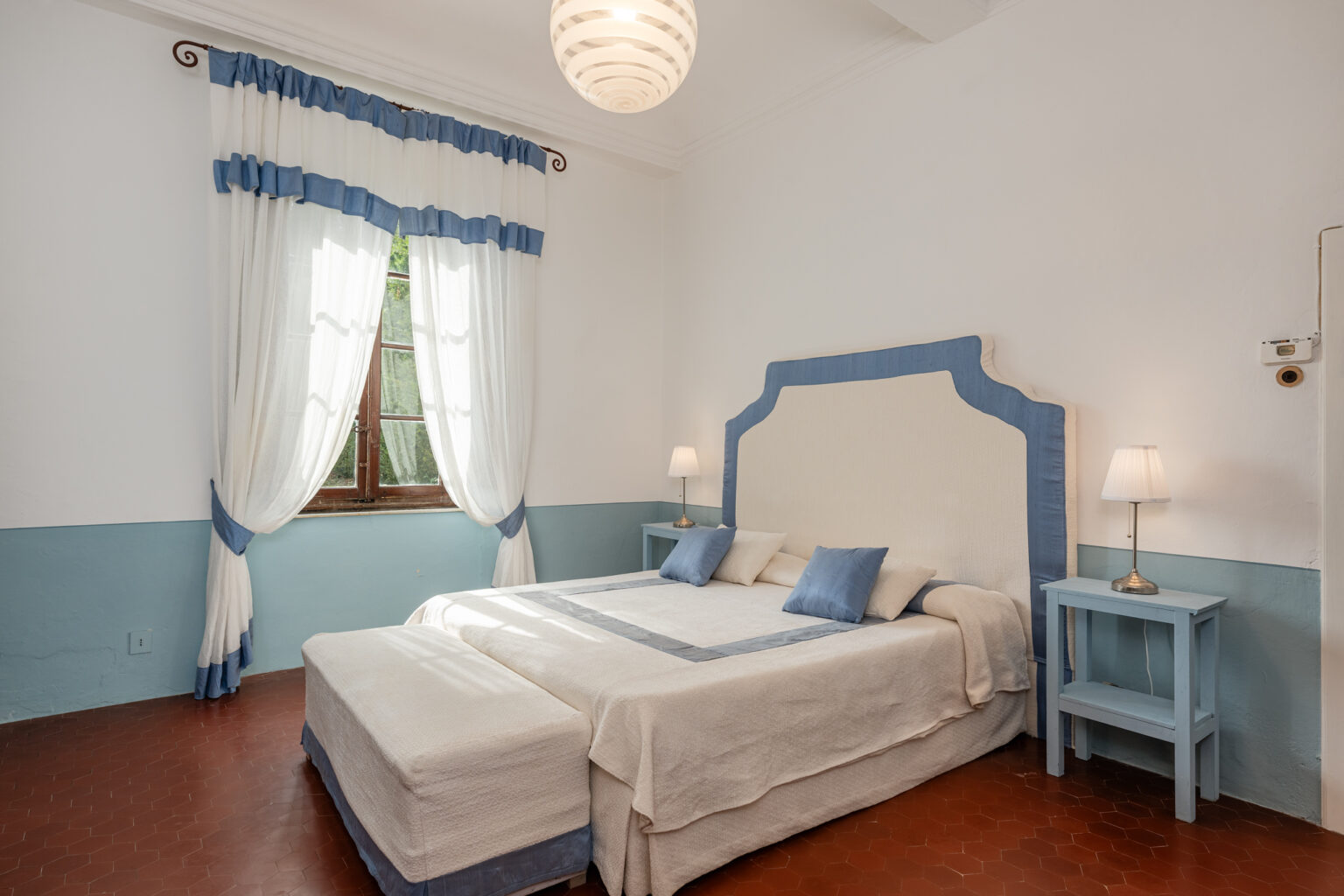 Cozy bedroom featuring a king-sized bed with blue accents, two bedside lamps, and elegant curtains, complemented by a light fixture and a window providing natural light. Ideal for a relaxing getaway. | Only on SopranoVillas