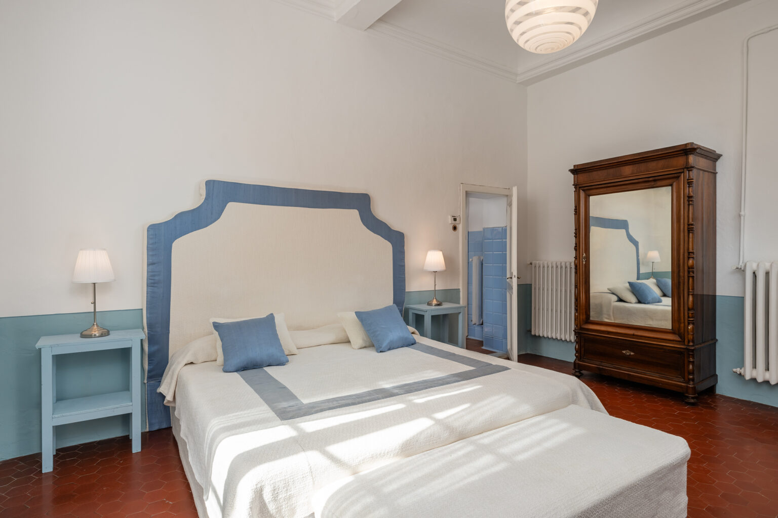 Bright and airy bedroom featuring a large bed with blue accents, two bedside lamps, a vintage wooden wardrobe, and a mirror, set against a clean white and light blue color scheme. | Only on SopranoVillas