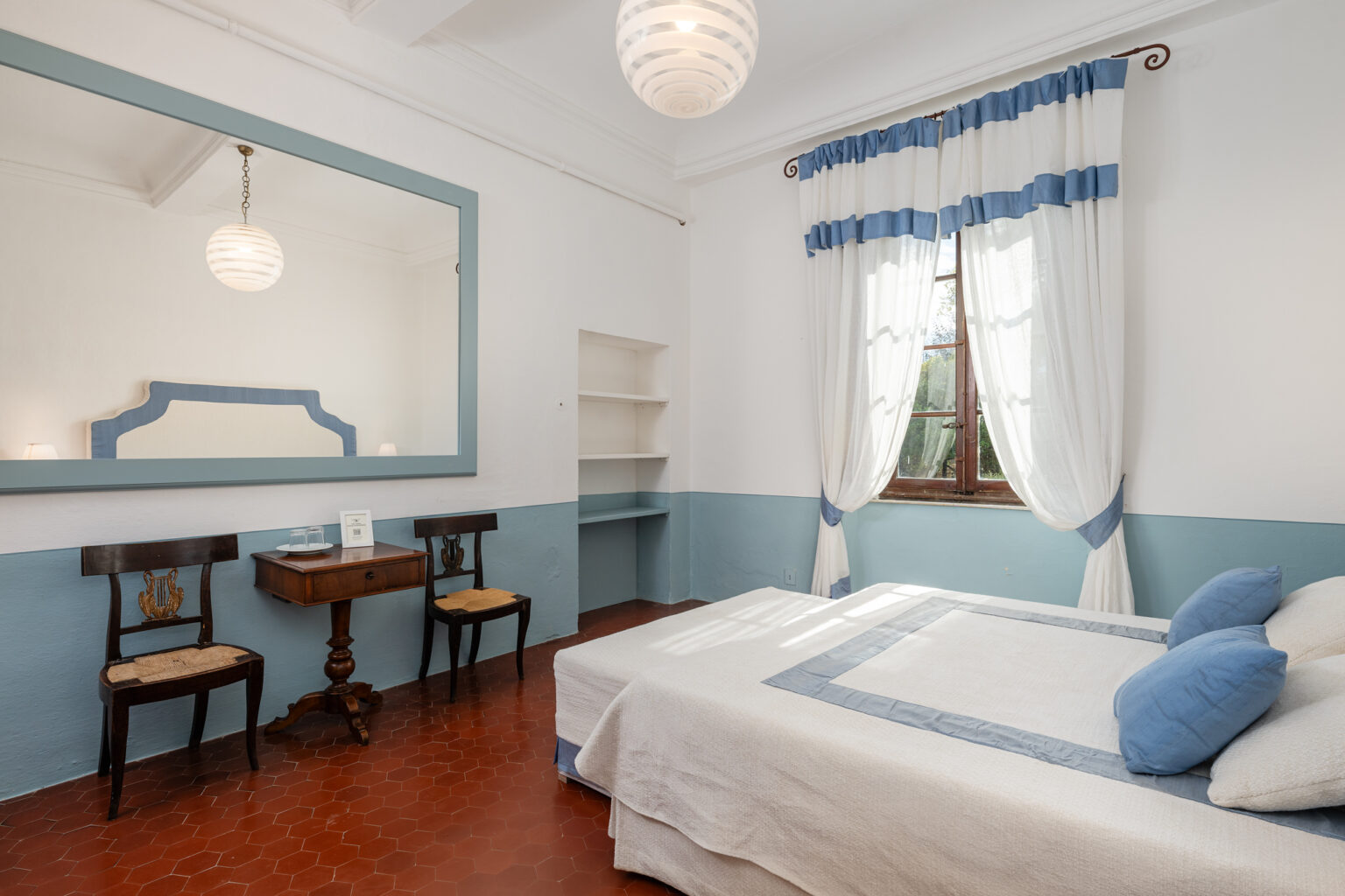 Bright and airy bedroom featuring a white and blue color scheme, a large mirror, a wooden table with two chairs, and a window with sheer curtains, creating a cozy and inviting atmosphere. | Only on SopranoVillas