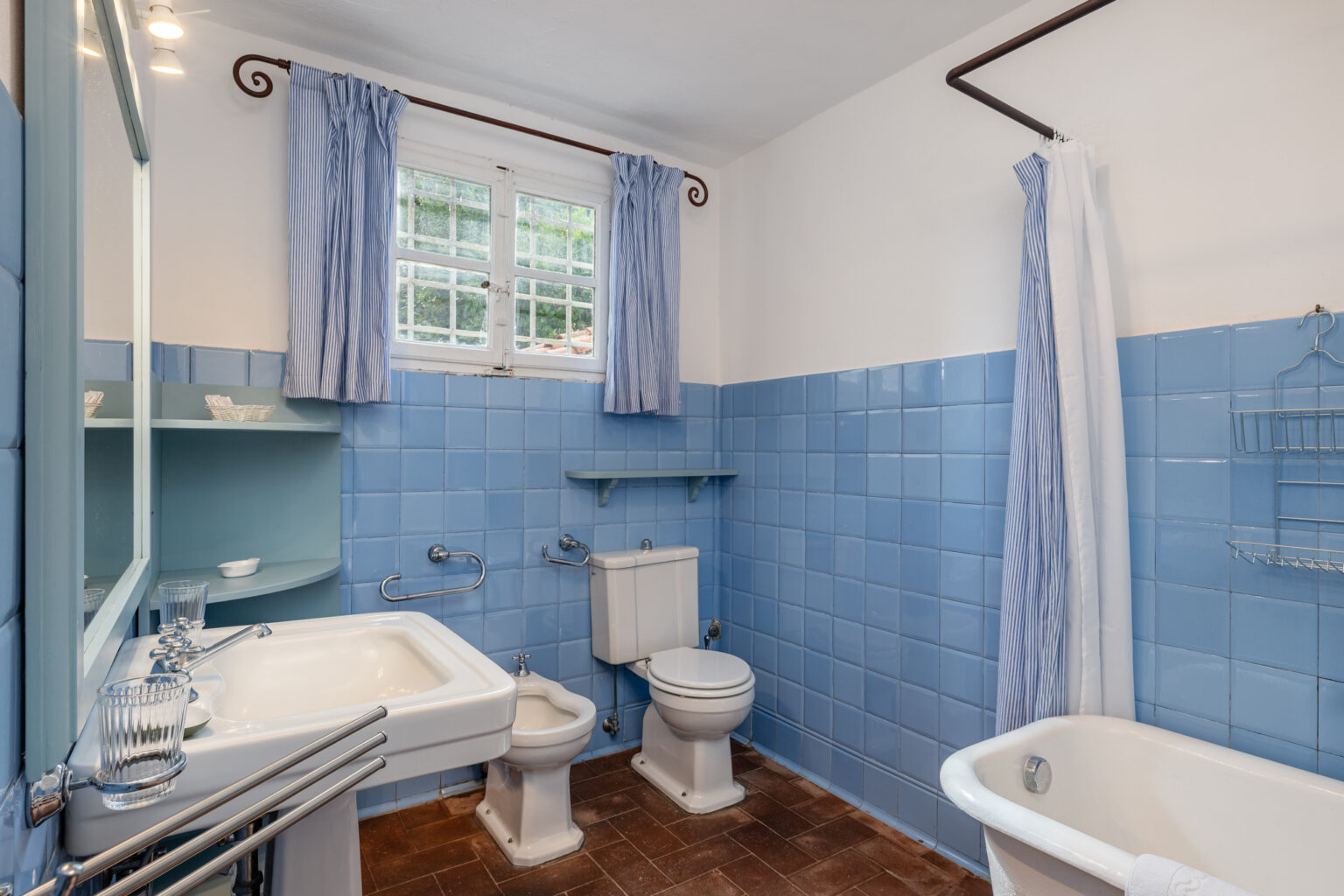 Bright and airy bathroom featuring blue tiled walls, a vintage bathtub, modern sink, and a toilet. Natural light filters in through a window adorned with striped curtains, enhancing the space's inviting atmosphere. | Only on SopranoVillas