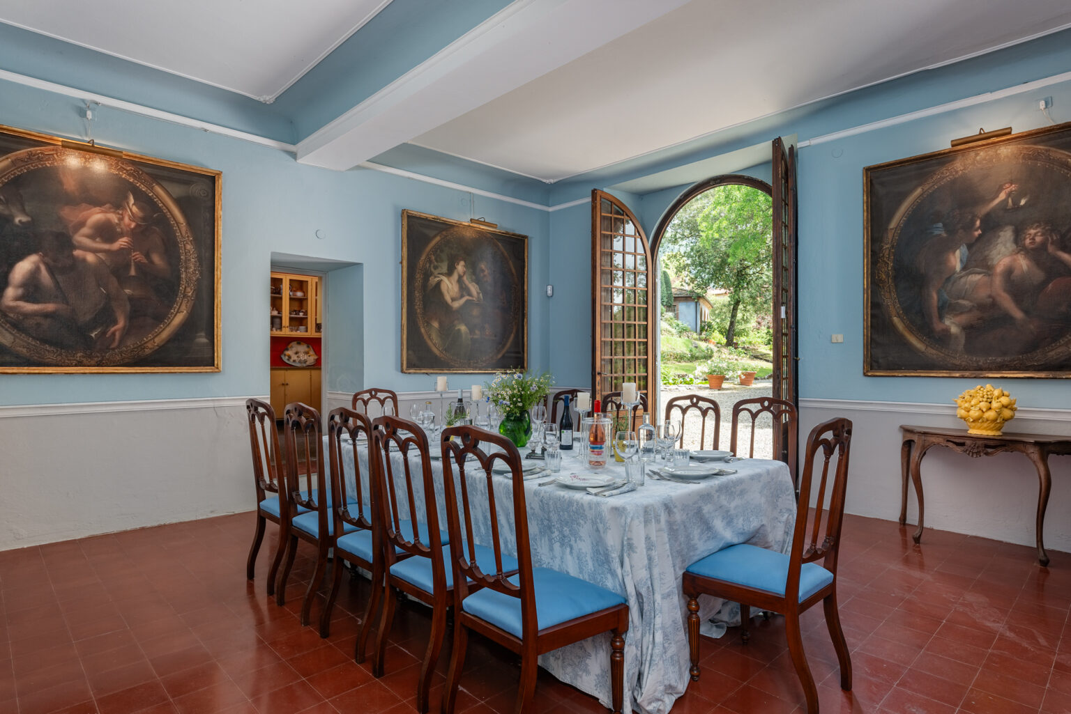 Elegant dining room with a blue color scheme, featuring a large table set for a meal, surrounded by wooden chairs. Two large paintings adorn the walls, and an open door reveals a lush garden outside, creating a serene atmosphere. | Only on SopranoVillas