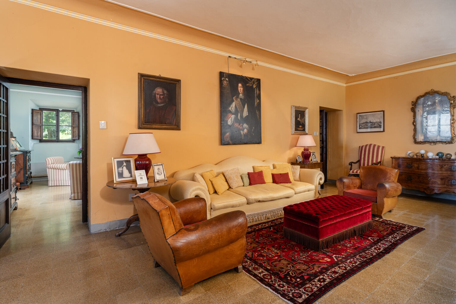 Cozy living room with warm orange walls, featuring a beige sofa, red velvet ottoman, and antique furniture. Decorated with framed portraits and a traditional rug, the space exudes a vintage charm. | Only on SopranoVillas