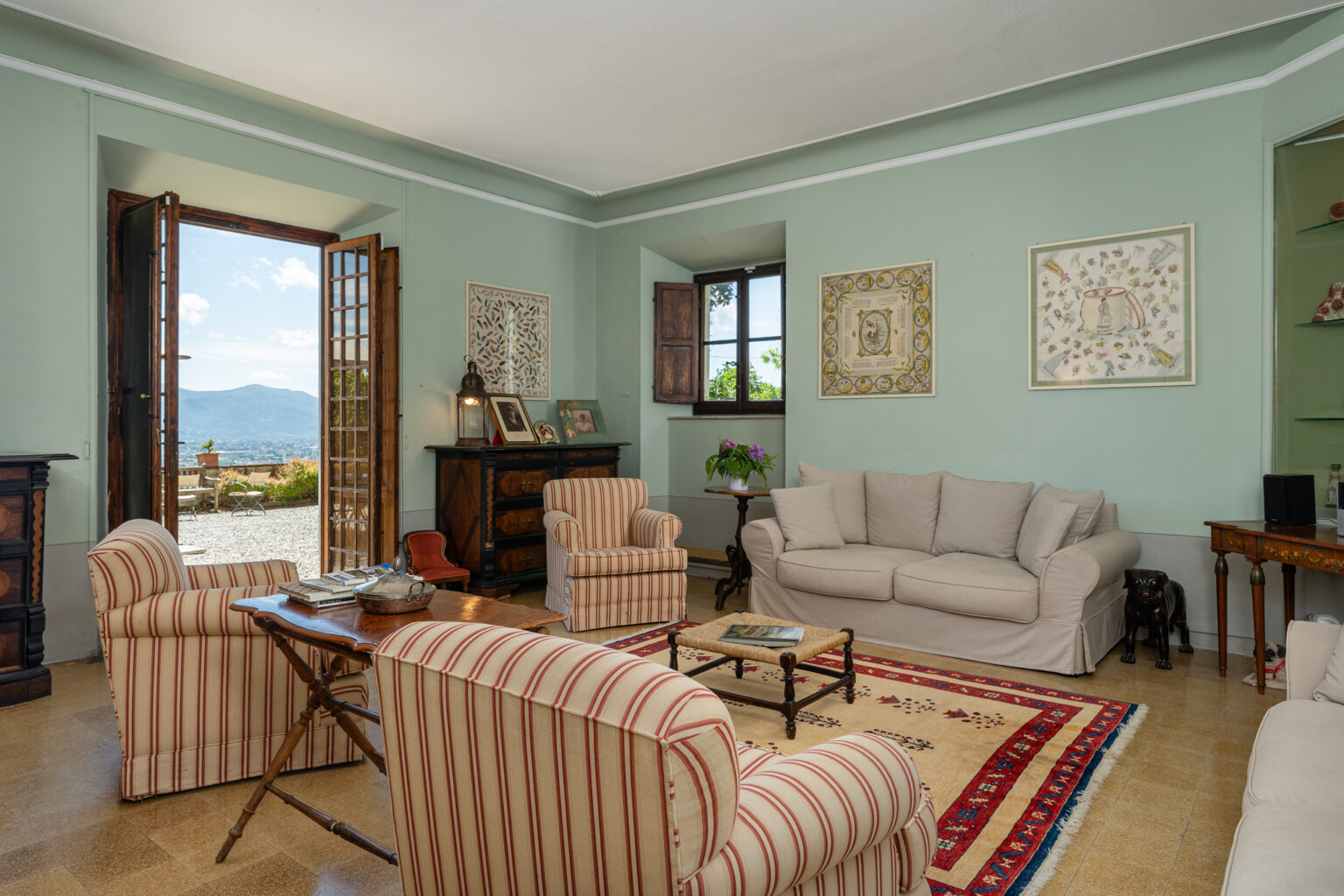 Cozy living room with a light green wall, featuring a beige sofa and striped armchairs, a wooden coffee table, and an open door leading to a scenic outdoor view. Decorated with artwork and plants, the space exudes a warm and inviting atmosphere. | Only on SopranoVillas